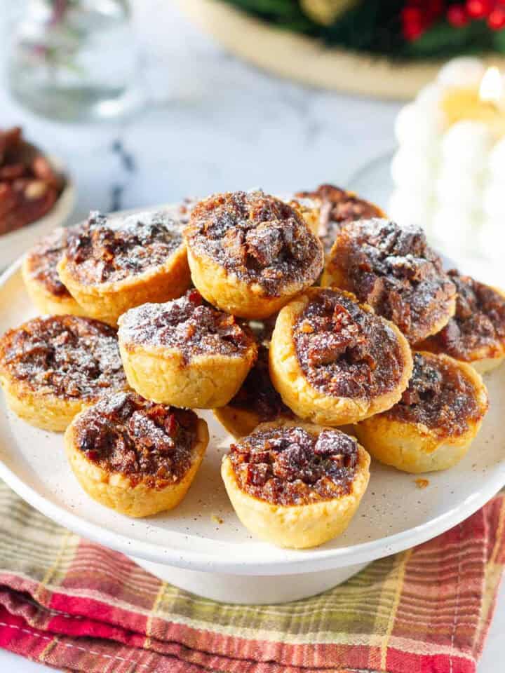 pecan tassies on a cake stand with a candle and pecans in the background.